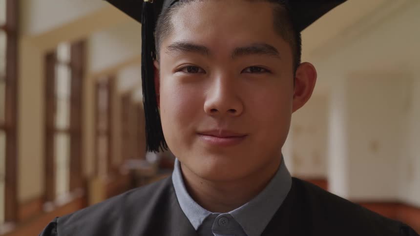 A young asian graduate standing in a hallway turns his head to smile into the camera