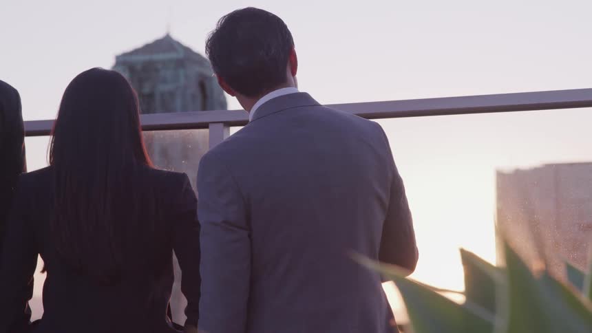 Asian businessman with a grey suit and businesswoman with a black suit stand on balcony and watch city skyline at sunset