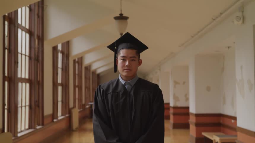 A young man dressed in his cap and gown for graduation stands inside a building