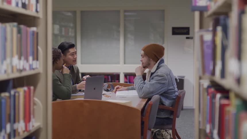 Three people sit together at a library table with a laptop