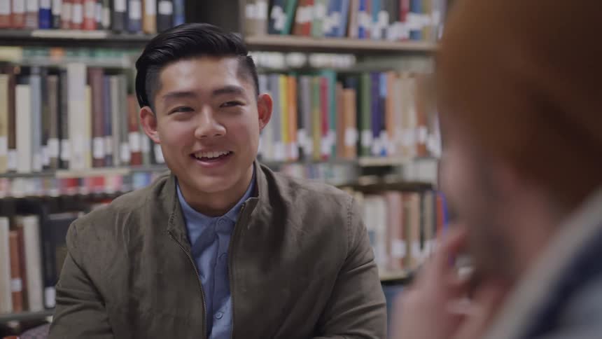 A young asian man talks to another man as they sit across from one another in a library