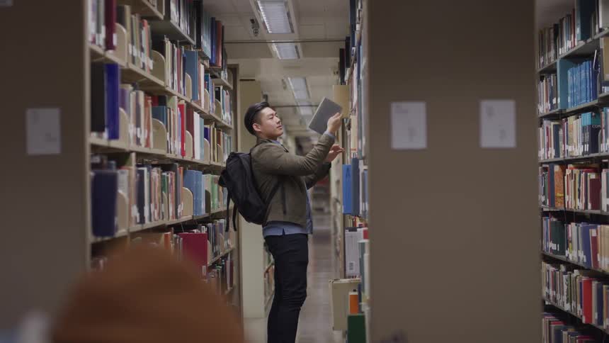 Teenage asian boy with a black backpack and pants searches for a book in the library