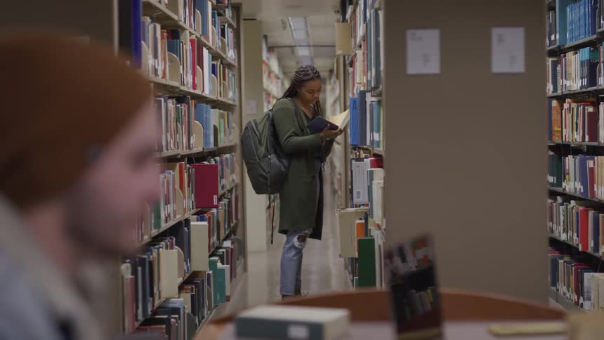 A woman with a backpack in a library taking a book from a shelf with a man with a hat in the foreground