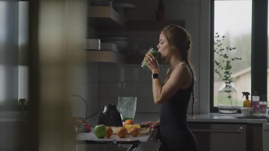 Woman with brown hair and sports black sleeveless top drinks a glass of smoothie in the kitchen