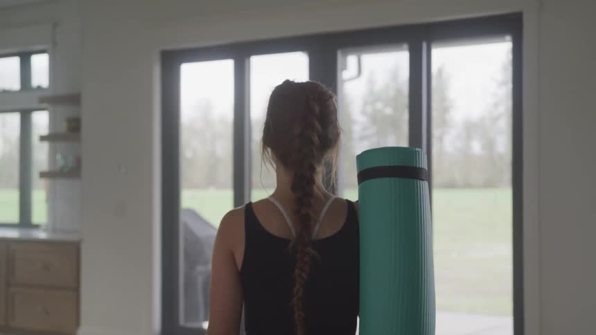 A woman carrying a yoga mat walks towards the door to leave her home