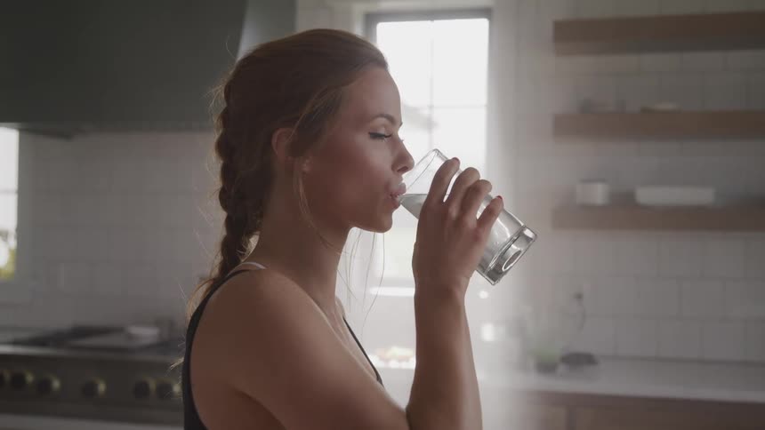A woman standing in a kitchen lifts a clear glass and drinks water