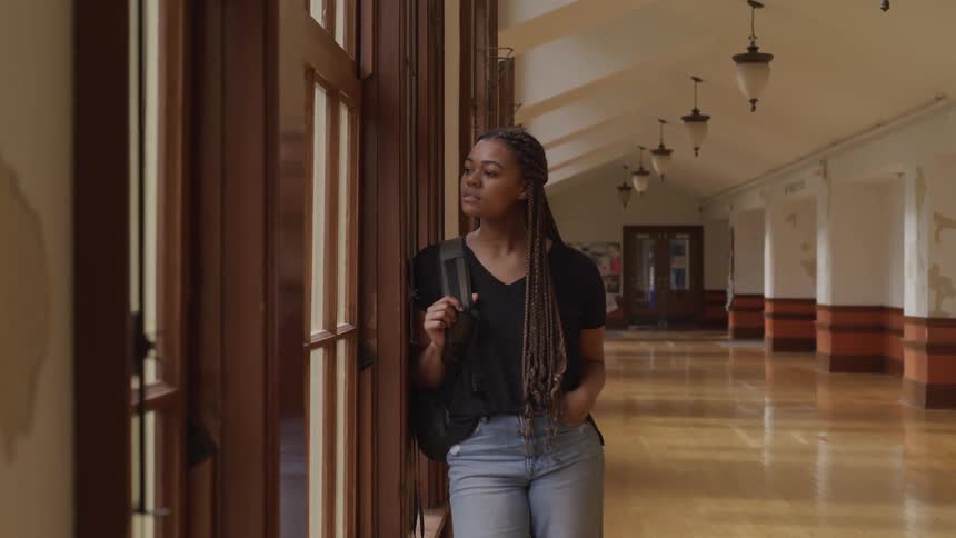 Black teenage girl with braided hair jeans and a backpack looks out the window at school corridor