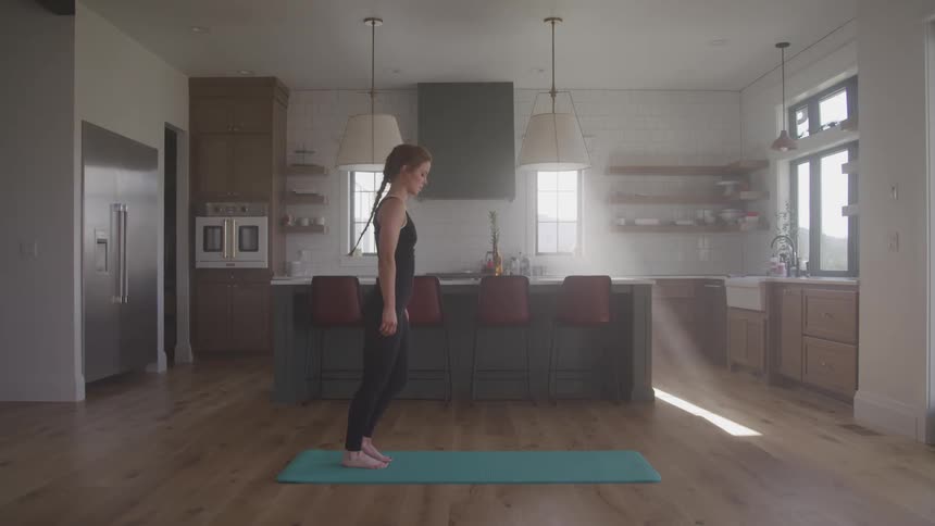 A woman stretches her leg forward in a yoga pose standing on a yoga mat in her kitchen