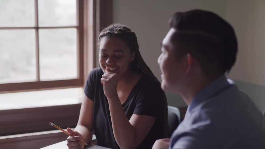 A pair of university students smile as they talk together while sitting at desks in a classroom