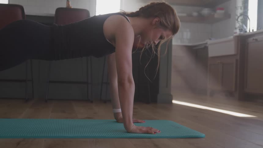 Woman does push ups on exercise mat on wooden floor near kitchen chairs