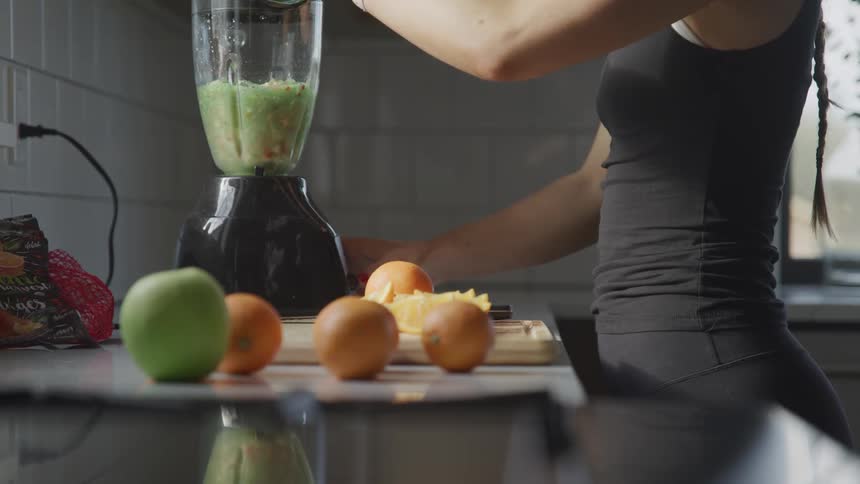 A woman wearing fitness apparel stands with her hand atop a blender as she prepares a blended fruit smoothie