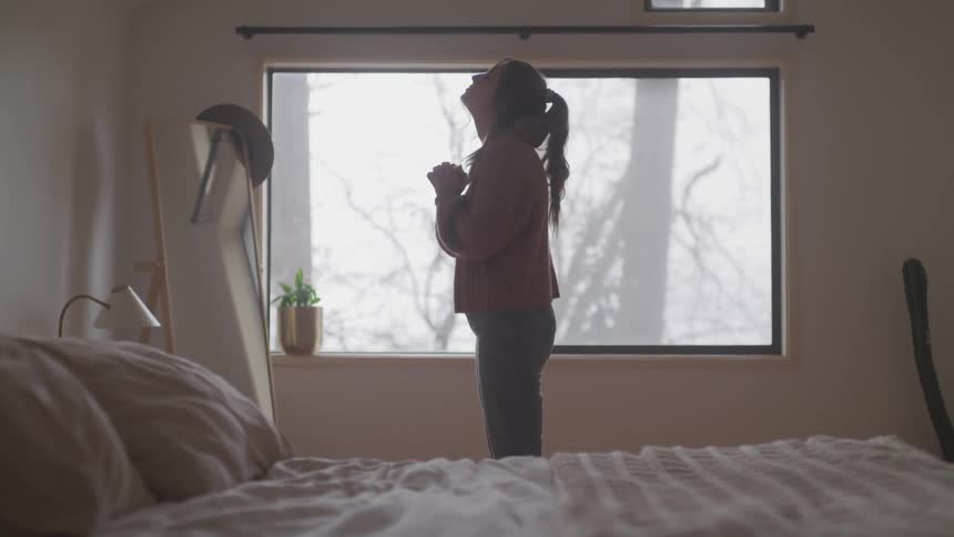 A woman clasps her hands together and prays while standing in her bedroom