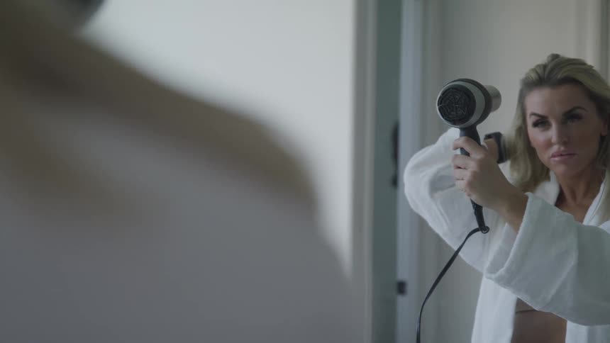 Woman with a white bathrobe dries dirt blonde hair using a hairdryer in bathroom