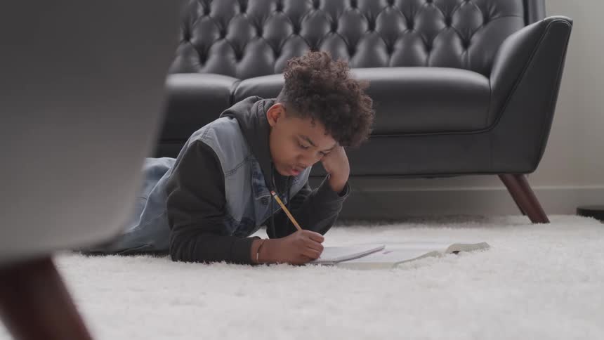 A boy does his homework while lying on the carpet in a living room