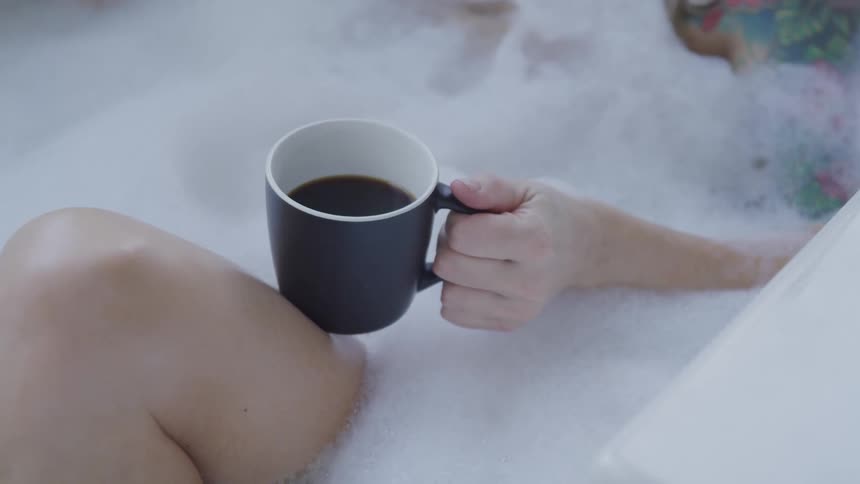 Woman takes a foamy bath in a bathtub holding a cup of black coffee in her hand