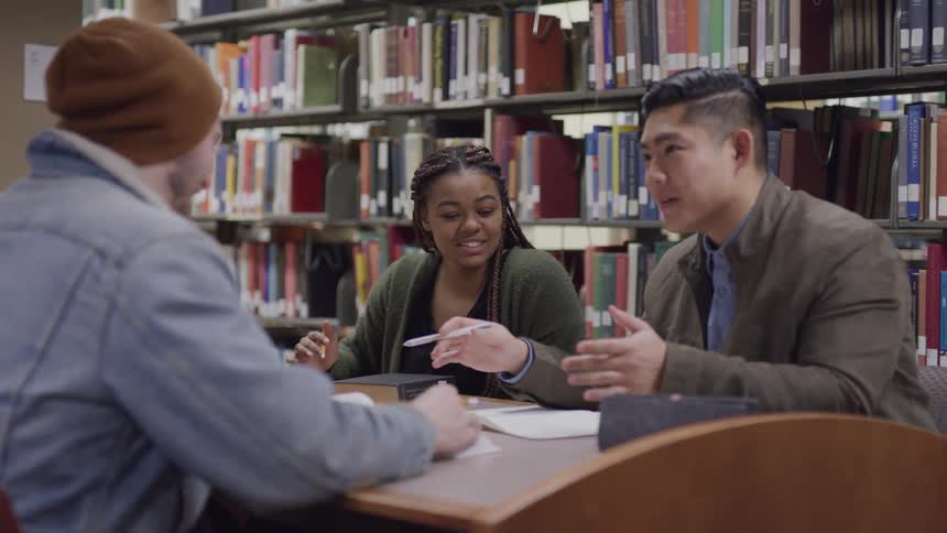Two men and a woman sitting at a table in a library with bookshelves and books in the background