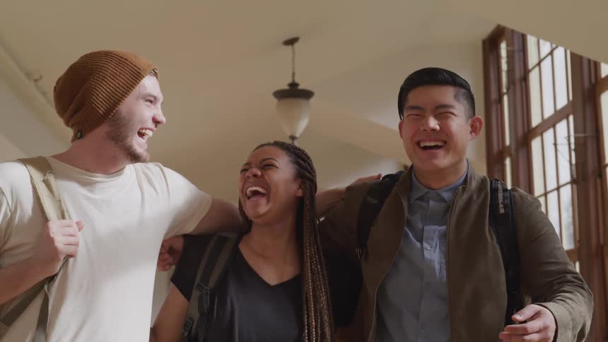 Students walk down university building hall laughing with their arms around each other