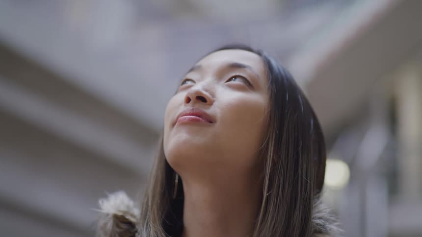 Asian girl with straight hair smiles slightly and looks up in a building