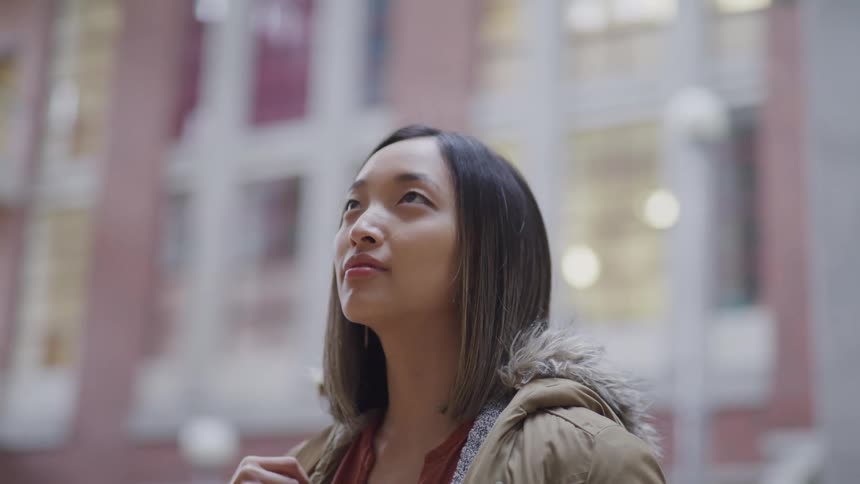 Teenage asian girl with straight hair and a brown coat looks up at buildings in the street