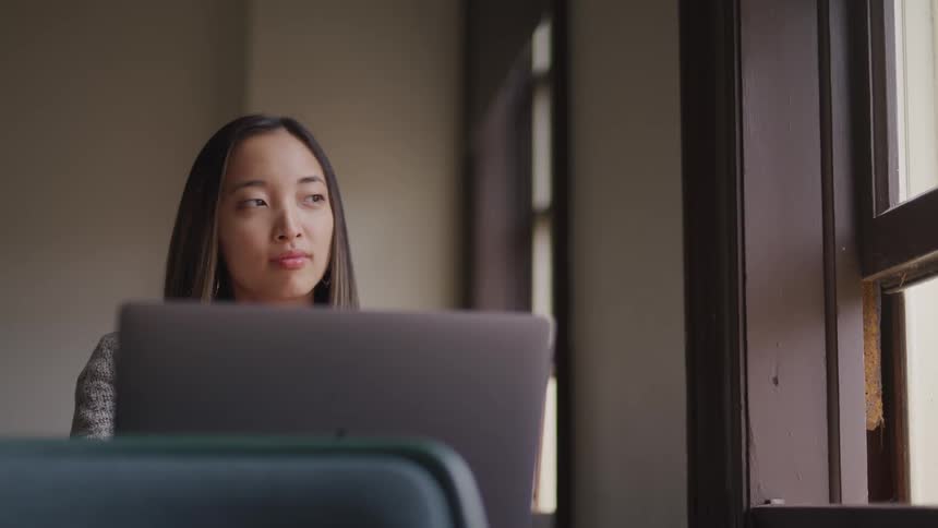 An asian female sitting at a table with a laptop inside a building