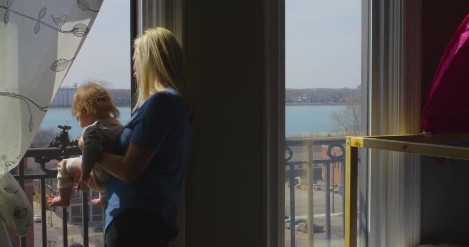 A woman holding a baby next to a window overlooking a body of water