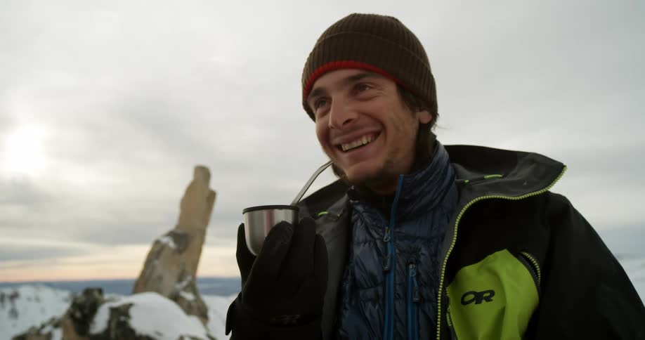 A man wearing winter gear holds a mate tea cup as he stands outdoors in the mountains
