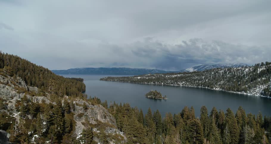 Pine trees and snow surround a mountain lake on a cloudy day