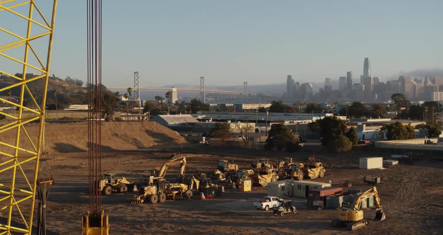 Crane towers over construction equipment near tree covered hill while fog obscures city skyline and nearby bridge