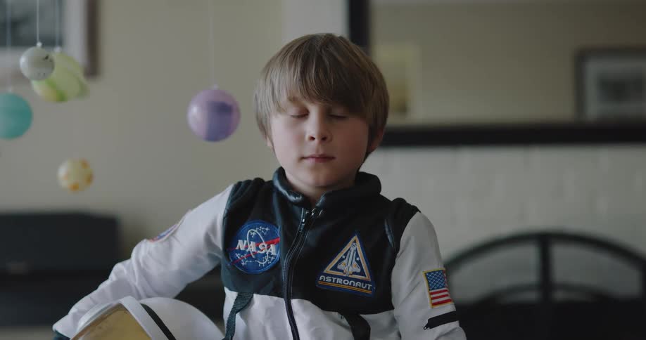 Young boy wearing an astronaut costume stands looking at the camera on the living room at home