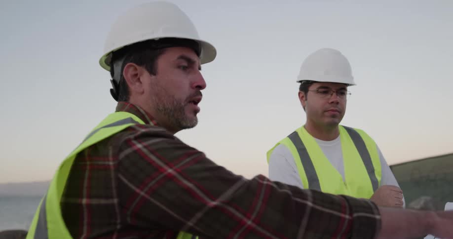 A man points over a worksite while looking at blueprints