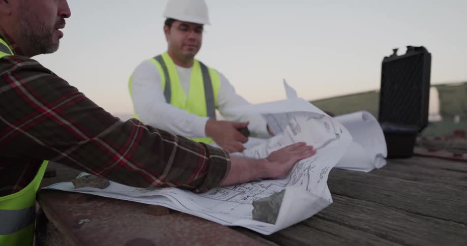 Two construction workers discuss building plans near ocean stopping to weight paper down with stones against wind