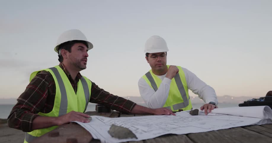 Two male construction workers talk to each other outside standing by a large bluepring
