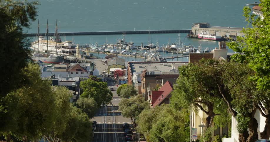 Two people walk across the street of a small town on the water that is filled with boats