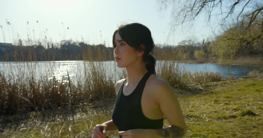 A woman jogs a trail near a pond with birds flying over the water surface on a sunny day