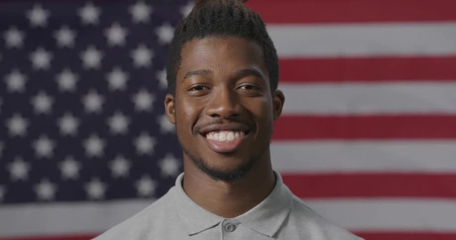 A young black man in front of the united states flag smiles for a portrait