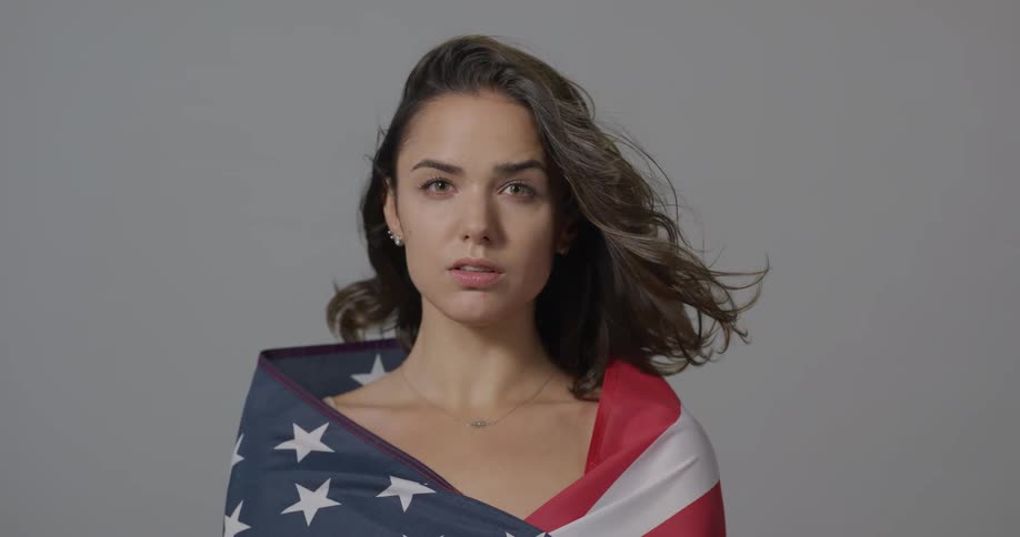 A naked young woman with an american flag draped around her shoulders looks into the camera as her hair blows in the wind