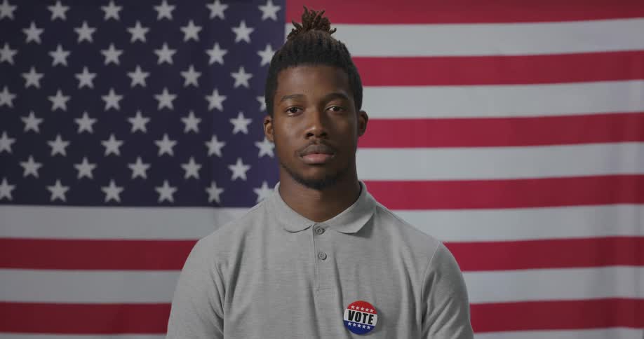Young man with a pin on his chest stands in front of an american flag looking at the camera