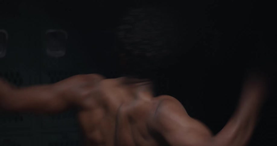 A teenage boy without a shirt on walks in to a locker room and throws punches in to the air and then lies down on a bench