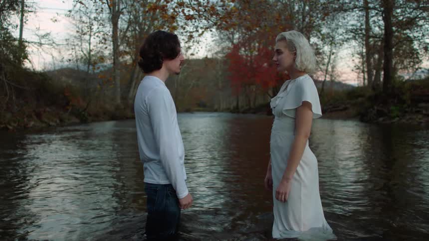 Man with a white sweatshirt and woman with a white dress stand in a river and look into each others eyes