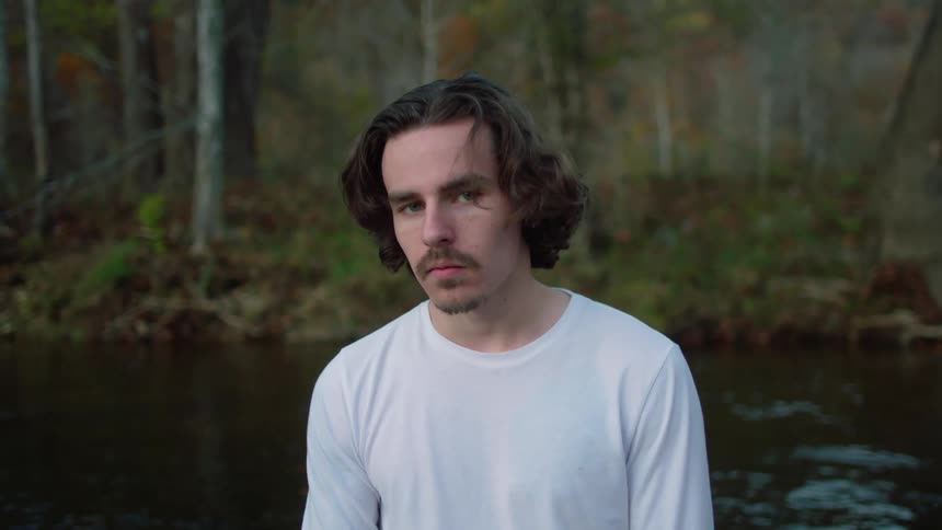 A man sitting at riverside in a woodland raises his head to look into the camera