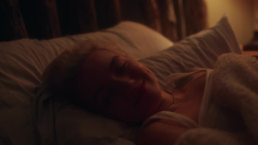 Blonde woman lies in bed and beams at the camera in bedroom