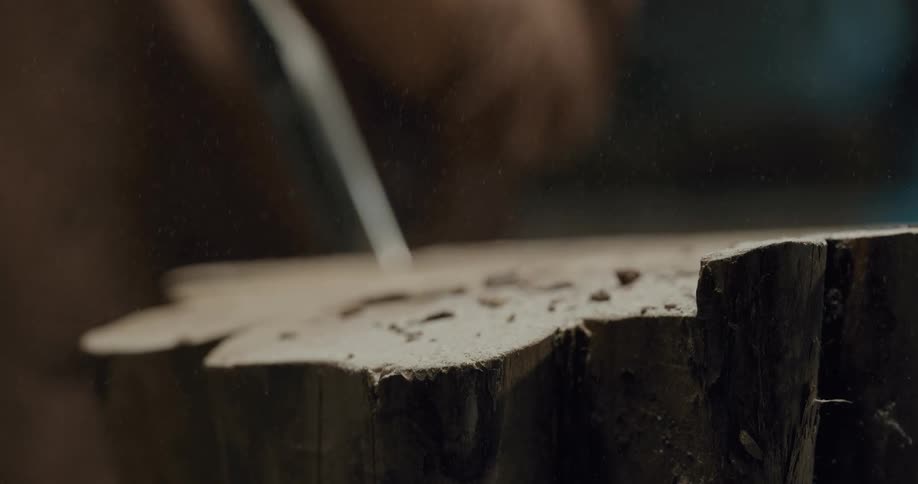 A man chips and carves wood and then blows the shavings off