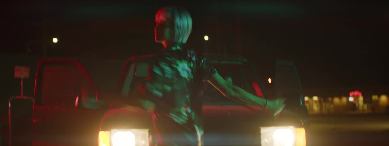 A woman dances wildly in the middle of the road at night in front of a vehicle with both doors open