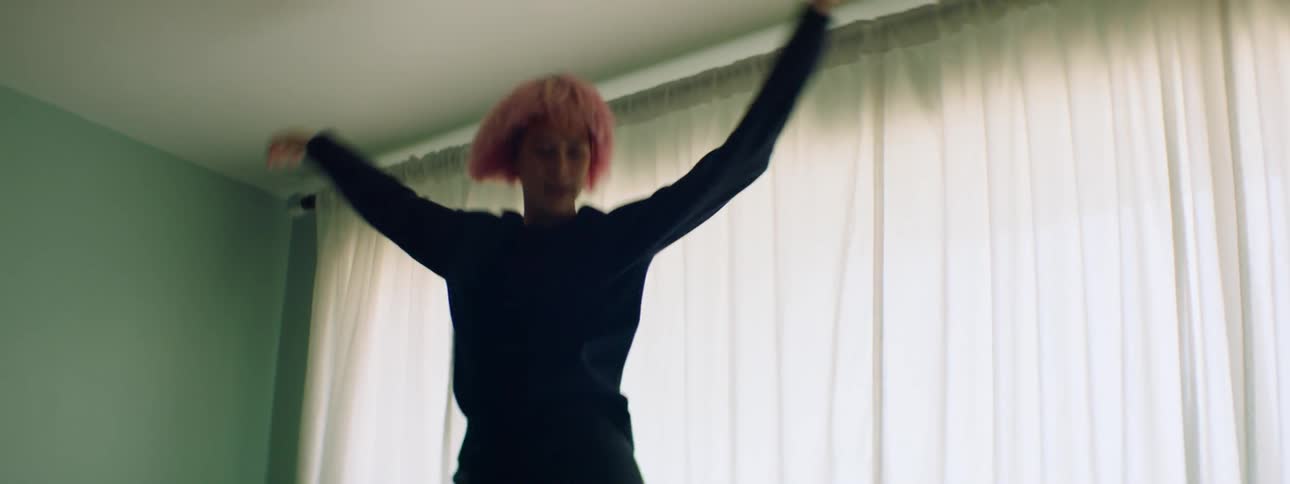 A young woman playfully dances atop a couch in front of a large window