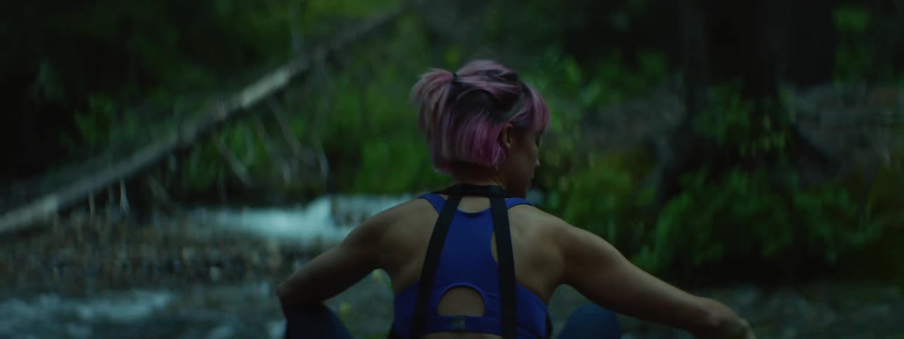 Woman with blue sports bras and pink hair sits on small river bank in the forest