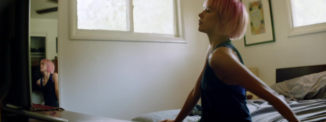 Young woman with pink hair dances while sitting on the edge of her bed