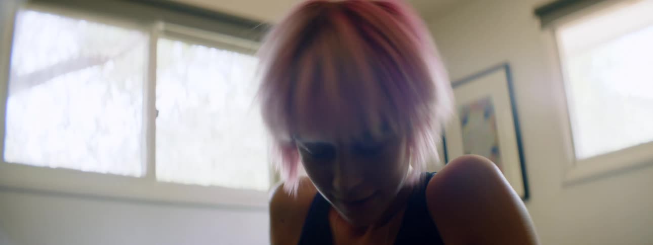 Woman with pink hair sits on a bed and dances wildly in a room
