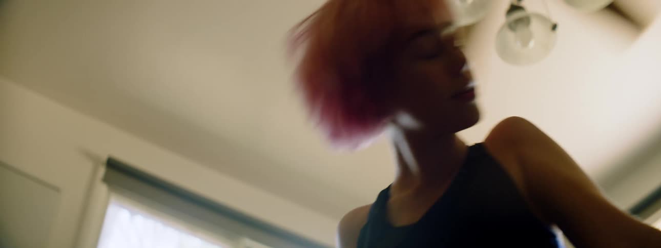 A woman with bright pink hair dances in her living room under a ceiling fan