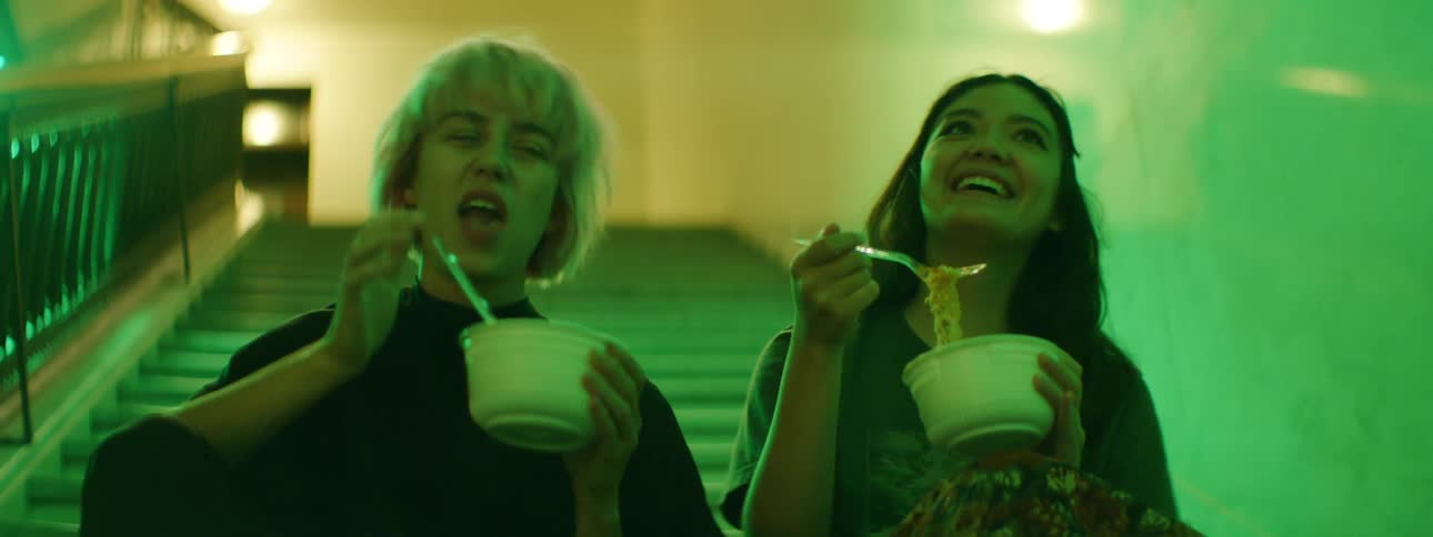 Two teenage girls sit in a stairwell eating noodles from a bowl