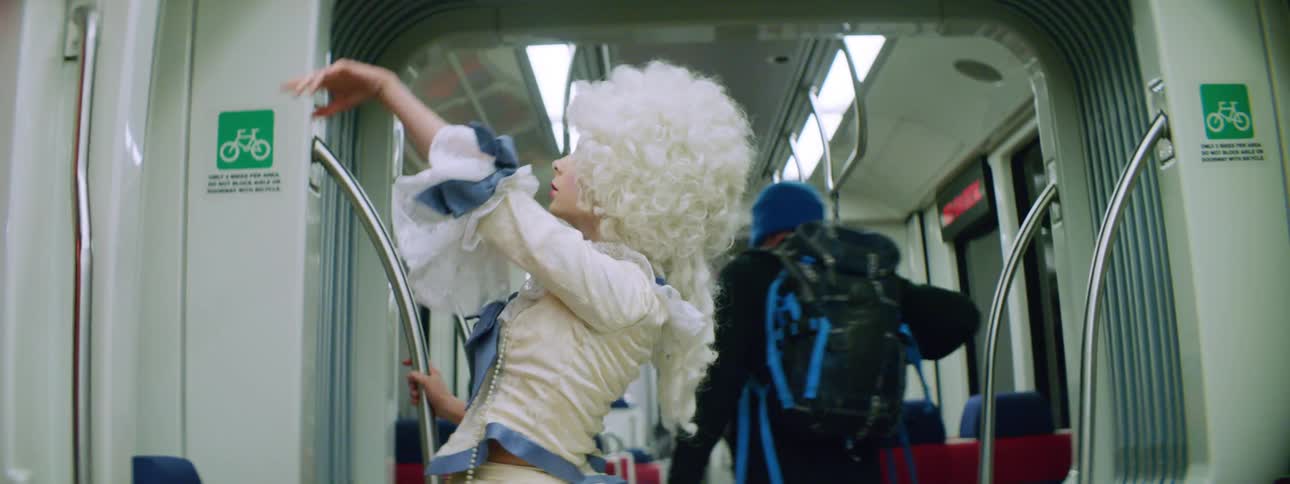 A young woman in a powdered wig and fancy victorian era dress dances strangely in a modern subway car